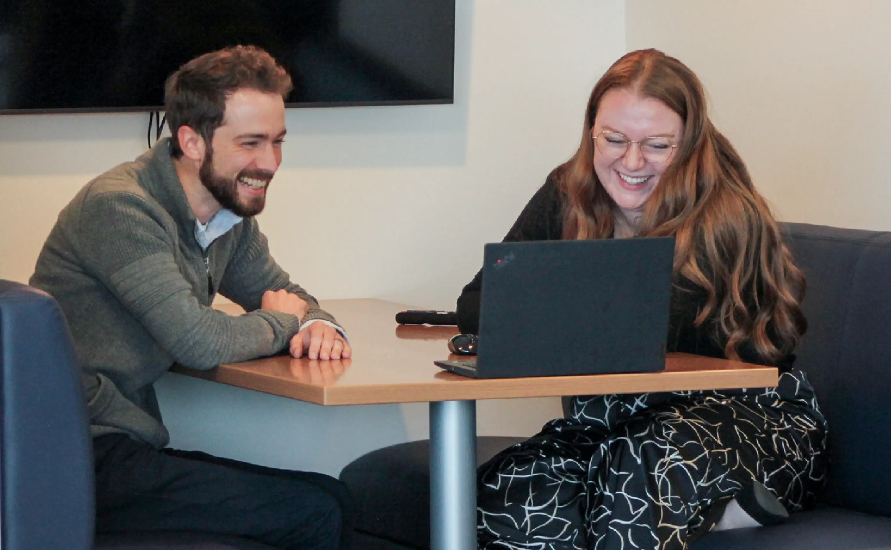 A man and a women sat at a desk looking at a laptop and laughing together
